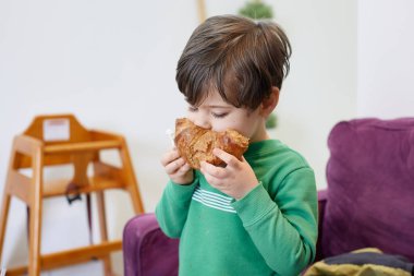 Yeşil kazaklı yakışıklı bir çocuk kanepede kruvasan yiyor.