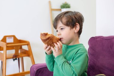 Yeşil kazaklı yakışıklı bir çocuk kanepede kruvasan yiyor.