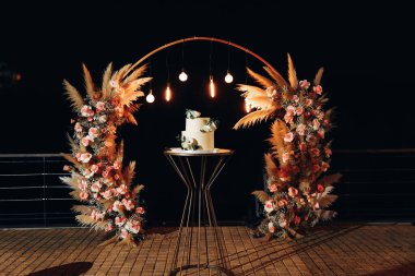 Çiçekli bir kemerin arka planında boho tarzında lüks bir düğün pastası. Akşam lambalarının altında düğün tatlısı. Düğün dekoru. Yüksek kalite fotoğraf