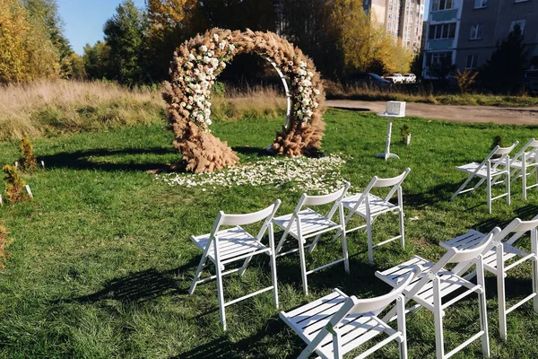 Wedding wooden arch in rustic style decorated with grass hay field color and flowers. Near wooden boxes with flower bouquets. Wooden arch and flower decoration.