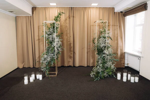 Indoor wedding ceremony with white wedding arch decorated with flowers and big white candles. High quality photo. High quality photo