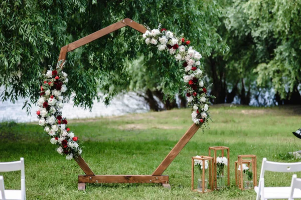 Wedding wooden arch in rustic style decorated with grass hay field color and flowers. Near wooden boxes with flower bouquets. Wooden arch and flower decoration.