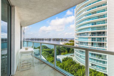 Balkondan Miami körfezine doğru güzel bir manzara, ağaçlı köprü, ufuk, mavi gökyüzü, tropikal bitki örtüsü, sandalyeli masa, turkuaz renkli camlar.