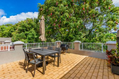 Masaları, sandalyeleri ve şemsiyesi olan büyük bir balkon, ızgara, beyaz korkuluk, arka planda yemyeşil ağaç, Fort Lauderdale 'de beyaz bulutlu mavi gökyüzü.