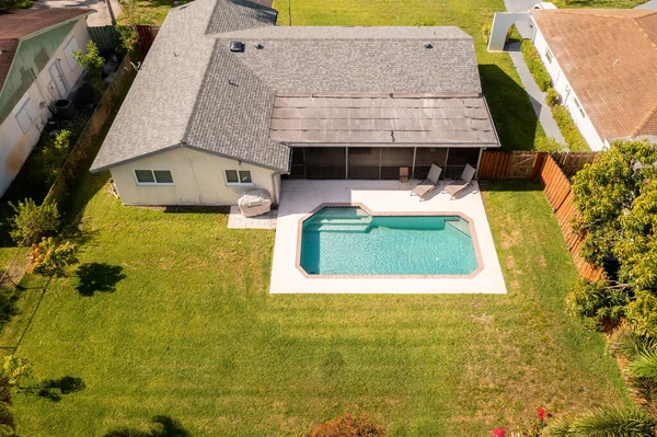 Aerial view from drone to backyard of house, swimming pool, covered patio with windows with mosquito netting, short grass, summer kitchen, sun loungers, wooden fence, trees and bushes, asphalt shingles