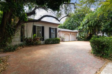 House facade in warm colors, 2 garage, cobblestone driveway, tropical vegetation, blue sky clipart