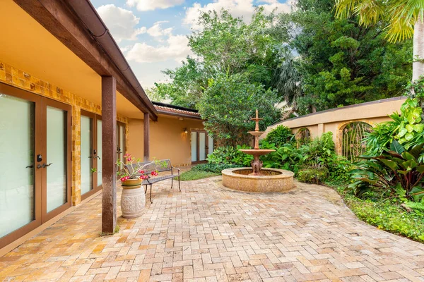 Backyard of elegant house in the Coral Brooks neighborhood, with abundant tropical vegetation, cobblestone floor, orange tones, fountain in the middle, blue sky in the background