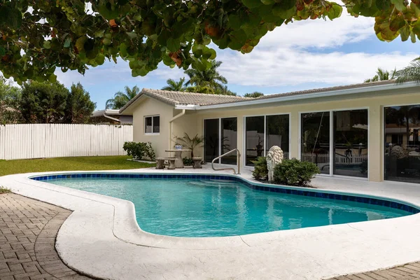 Backyard with pool with brick deck, covered patio, sun loungers, short grass, tropical tree, white wooden fence,