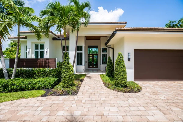 Facade of a beautiful place, with a front garden made up of palms, short grass and tropical plants, in Coral Ridge in Miami, driveway, sidewalk and street