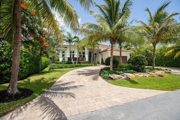 Facade of a beautiful place, with a front garden made up of palms, short grass and tropical plants, in Coral Ridge in Miami, driveway, sidewalk and street