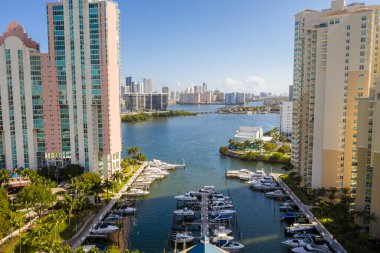 Arka planda şehir, mavi gökyüzü, ağaçlı ve palmiyeli tropikal bitki örtüsü, körfez tekneli iskele, yatlı insansız hava aracı görüntüsü.