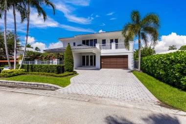 Miami 'nin Riviera Adaları' ndaki zarif modern evin ön cephesi, özel duvar, garaj, balkon, siyah fayanslar, tropikal bitki örtüsü ve kısa çimen, beyaz duvarlar, mavi gökyüzü.