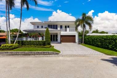 Miami 'nin Riviera Adaları' ndaki zarif modern evin ön cephesi, özel duvar, garaj, balkon, siyah fayanslar, tropikal bitki örtüsü ve kısa çimen, beyaz duvarlar, mavi gökyüzü.