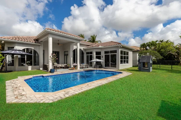 Backyard of elegant house with swimming pool, short grass, outdoor furniture, umbrella, house with gray walls and white details, roof with red tiles, blue sky with clouds
