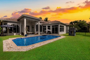 Backyard of elegant house with swimming pool, short grass, outdoor furniture, umbrella, house with gray walls and white details, roof with red tiles, blue sky with clouds clipart