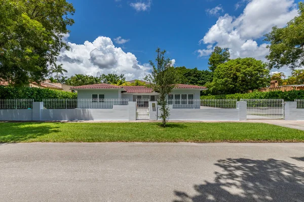 Entrance of a gray house with red tiles, in the Granada neighborhood, in Coral Gables, Miami, trees, blue sky, driveway, footpath, fence with white bars