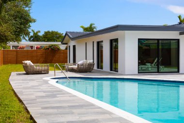 Miami 'nin Highland Lakes mahallesindeki zarif evin arka bahçesi, kısa çimenler, beyaz duvarlar, aynalı pencereler, yüzme havuzu, kaldırım, ahşap çitler, siyah fayanslar, mavi gökyüzü, palmiyeler, güneş pansiyonları.
