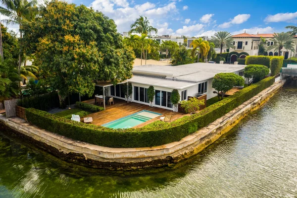 Beautiful patio with dark wood floor, swimming pool, bushes, trees, wooden coffee table lounge chairs, blue sky with clouds in Biscayne Point
