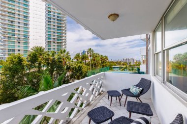 Palmiye ağaçlarıyla balkondan manzara, turkuaz pencereli beyaz binalar, tenis kortu, arka planda bulutlu binalar.