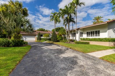 Koloni tarzı ev cephesi, kırmızı fayanslarla çevrili, tropikal bitki örtüsü, garaj yolu ve ön kapıya giden yol, mavi gökyüzü