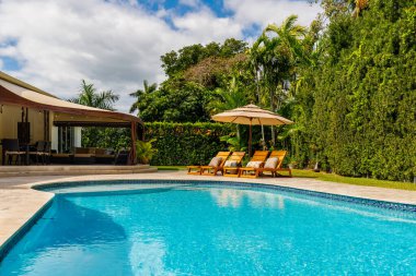 Havuzu olan güzel bir avlu, şemsiyeli güneşli şezlonglar, açık mobilyalarla kaplı avlu, özel duvar, palmiyeler, kısa çimenler, mavi gökyüzü