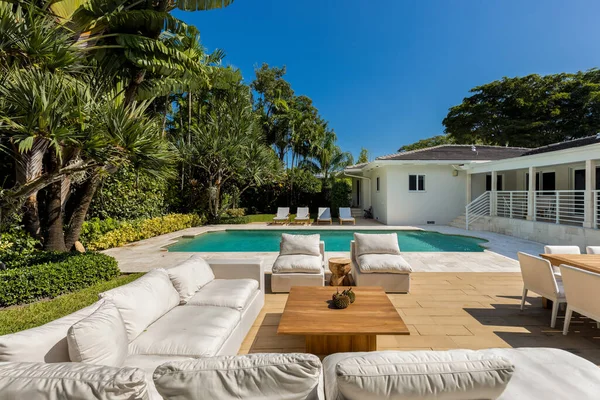Backyard of elegant modern house, with swimming pool, outdoor furniture, short grass, wooden and tile floor, privet wall, tropical plants, palms, bushes, blue sky