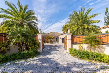 Ahşap kapılı, palmiyeli, çalılıklı, mavi gökyüzü olan bej renkli koloni tarzı bir evin ön cephesi.