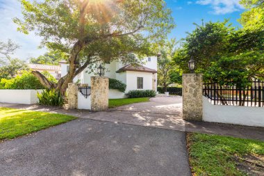 Elegant facade of a colonial-style house in the Riviera neighborhood, in Coral Gables, white walls with red tiles, stone details, short grass, trees, driveway and path to the front door clipart