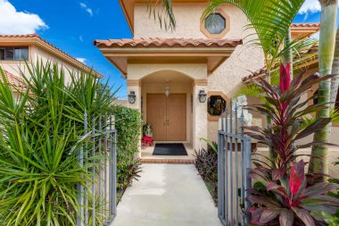 Kırmızı fayanslı, kaldırımlı, caddeli, garajlı, ön kapıya giden yol, kısa çimenler, palmiyeler, çalılar, tropikal bitkiler, mavi gökyüzü