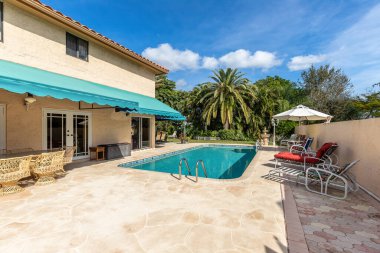 Kırmızı kiremitli bej renkli koloni tarzı evin arka bahçesi, turkuaz her şey, yüzme havuzu, açık mobilyalar, heykeller, fıskiye, fener, yaz mutfağı, kısa çimenler, palmiyeler, çalılar, tropikal bitkiler, mavi gökyüzü