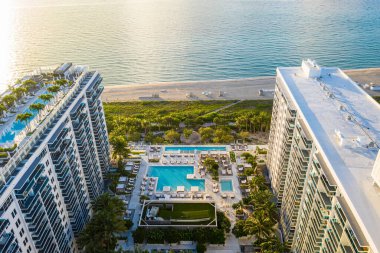 Aerial drone shot of Miami Beach coastline with towers and modern buildings, blue sky, bay, waves, beach, tropical vegetation, swimming pools clipart