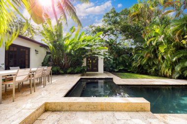 Havuzu, kaplıcası ve şelalesi olan güzel bir bahçe, kaldırım taşı zemini, kısa çimenleri, palmiye duvarları, bol tropikal bitki örtüsü, arka planda sandalye, masa ve şezlonglar ve mavi gökyüzü gibi açık hava mobilyaları.