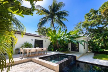 Havuzu, kaplıcası ve şelalesi olan güzel bir bahçe, kaldırım taşı zemini, kısa çimenleri, palmiye duvarları, bol tropikal bitki örtüsü, arka planda sandalye, masa ve şezlonglar ve mavi gökyüzü gibi açık hava mobilyaları.