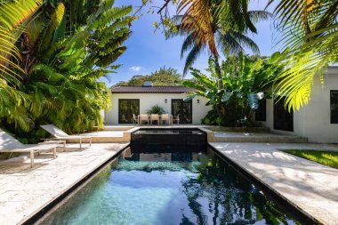 Havuzu, kaplıcası ve şelalesi olan güzel bir bahçe, kaldırım taşı zemini, kısa çimenleri, palmiye duvarları, bol tropikal bitki örtüsü, arka planda sandalye, masa ve şezlonglar ve mavi gökyüzü gibi açık hava mobilyaları.