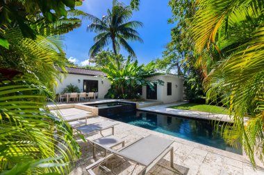 Havuzu, kaplıcası ve şelalesi olan güzel bir bahçe, kaldırım taşı zemini, kısa çimenleri, palmiye duvarları, bol tropikal bitki örtüsü, arka planda sandalye, masa ve şezlonglar ve mavi gökyüzü gibi açık hava mobilyaları.