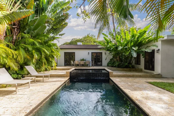 Beautiful garden with pool with spa and waterfall, with cobblestone floor, short grass, wall of palms, abundant tropical vegetation, outdoor furniture such as chairs, tables and loungers and blue sky in the background