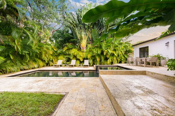 Beautiful garden with pool with spa and waterfall, with cobblestone floor, short grass, wall of palms, abundant tropical vegetation, outdoor furniture such as chairs, tables and loungers and blue sky in the background