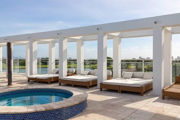 View from a terrace in the Bay Harbor Island neighborhood in Miami-Dade, outdoor beds to rest and relax in front of a swimming pool, golf course, blue sky in the background