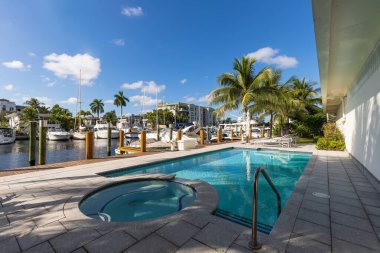 View from the pier in the Nurmi Isles neighborhood in Fort Lauderdale, canals with boats everywhere, swimming pool, sun loungers, houses and modern buildings, tropical climate with palms, bushes, trees, blue sky and skyline on the sea