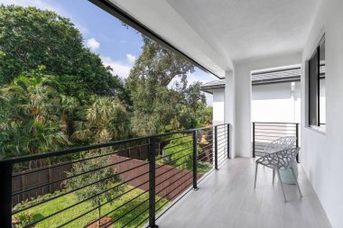 View from balcony of fenced backyard with wooden fences, short grass and tree in the middle, tropical vegetation around, outdoor chairs in gallery, in the Tarpon River neighborhood of the city of Fort Lauderdale