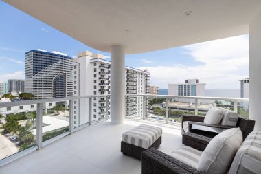 View of elegant balcony with wooden and wicker furniture in opaque colors, glass railing, with urban landscape and sea in the background,