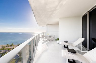 Shot of white balcony with view of fort lauderdale beach in Central Beach neighborhood, outdoor furniture, chairs and table, palms and tropical vegetation, turquoise sea and blue sky