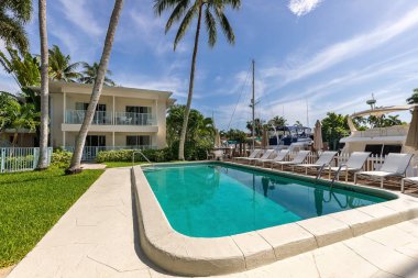 Point of view of pool, with parked boats, sun loungers with umbrellas, white metal fence fence, palms, balls, blue sky, in the Nurmi Isles neighborhood, Fort Lauderdale, Florida
