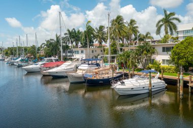Aerial drone view of wooden decked pier, with parked boats, short grass, tropical vegetation, palm trees, luxury houses and condos, blue sky, in Nurmi Isles neighborhood, Fort Lauderdale, Florida