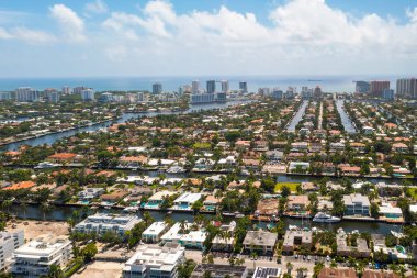 Aerial drone view of the Nurmi Isles neighborhood in Fort Lauderdale, canals with boats and piers around, tropical plants, palm trees, modern buildings and houses, sea on the horizon, blue sky