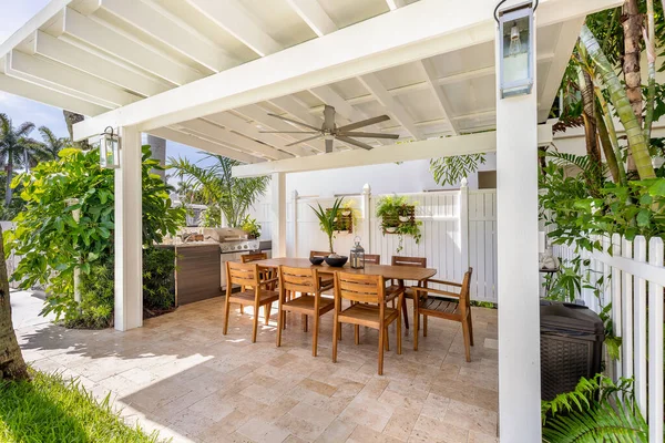 Beautiful outdoor kitchen under covered patio with outdoor tables and chairs, tropical plants hanging and in flower beds, white wooden fence, ceiling fan, palms and blue sky