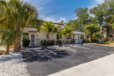 Facade of multipurpose building located in Fort Lauderdale, parking area, tropical vegetation, doors, windows, gray and white tones, trees, palms, sidewalks and stone beds, and short grass