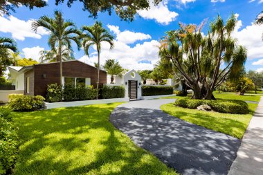 Beautiful facade of a luxury mansion located in Pinecrest, Miami-Dade with a vine on the wall, surrounded by trees and palms, short grass, driveway, front door, green, street and blue sky