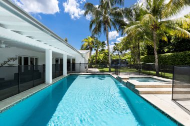 Backyard of home with fenced pool and spa, park with short grass, veranda with outdoor furniture, palms, blue sky
