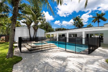 Backyard of home with fenced pool and spa, park with short grass, veranda with outdoor furniture, palms, blue sky